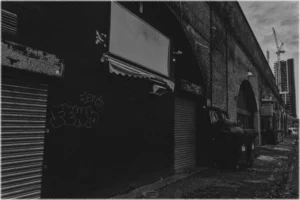 Eine körnige Schwarz-Weiß-Aufnahme einer rauen, städtischen Szenerie. Links dominiert eine dunkle Gebäudefassade mit heruntergelassenem Rollladen, Graffiti und einem leeren, weißen Schild über einer kleinen Markise. Rechts erstreckt sich eine lange Backsteinmauer mit Bögen (im Stil eines Eisenbahnviadukts), vor der mehrere große, schwarze Müllcontainer aufgereiht sind. Im Hintergrund ragen ein Baukran und ein modernes Hochhaus in den bewölkten Himmel.