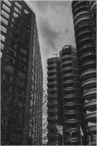 Eine körnige Schwarz-Weiß-Aufnahme aus der Froschperspektive, die steil nach oben auf moderne Hochhausarchitektur blickt. Links ragt ein massives, rechtwinkliges Gebäude mit einer strengen Rasterfassade in den Himmel. Rechts stehen im Kontrast dazu hohe, zylindrische Türme mit geschwungenen, horizontalen Balkonbändern. Im unteren Bildbereich sind die dunklen Silhouetten kahler Baumäste sowie drei Fahnenmasten (einer mit Union Jack) zu erkennen, die vor dem grauen, bewölkten Himmel stehen.