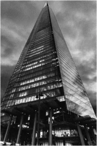 Eine körnige Schwarz-Weiß-Aufnahme, die in steiler Froschperspektive an der Fassade des „The Shard“ hinaufblickt. Die beleuchteten Fensterreihen bilden ein geometrisches Raster, das sich zur Spitze des Turms hin verjüngt, während das Gebäude kontrastreich vor einem düsteren, wolkenverhangenen Himmel aufragt.