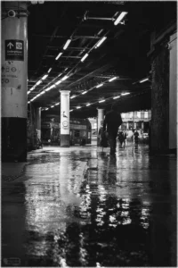 Eine atmosphärische Schwarz-Weiß-Aufnahme in einer nassen, städtischen Unterführung. Der Vordergrund wird von dunklem, regennassem Asphalt dominiert, auf dem sich die Lichter der Deckenbeleuchtung stark spiegeln. Eine einzelne Person geht als dunkle Silhouette von der Kamera weg in den Tunnel hinein. Auf der linken Seite steht ein massiver Pfeiler mit einem Wegweiser zur „Underground“, und im Hintergrund ist schemenhaft ein Doppeldeckerbus zu erkennen.